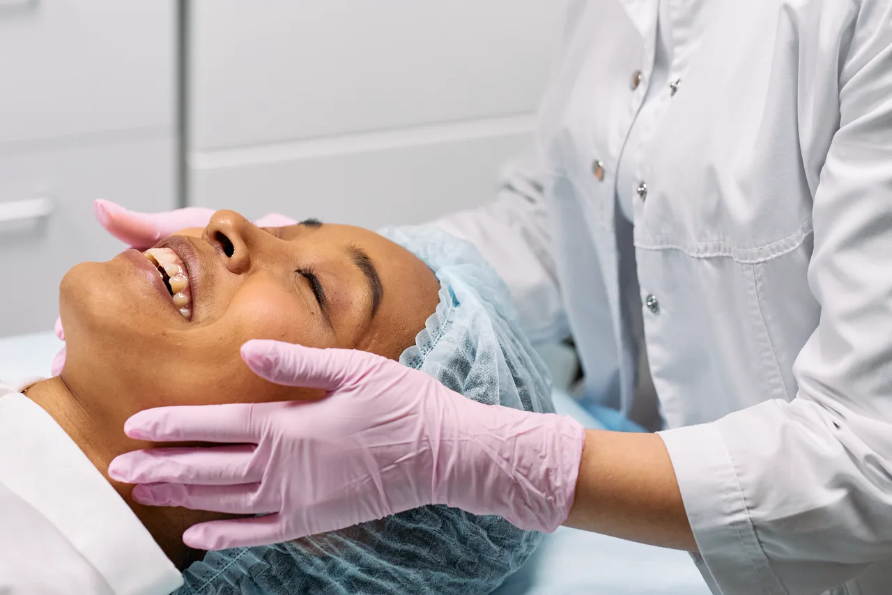 A smiling client lying back before a skin treatment, a practitioner in pink gloves beside her