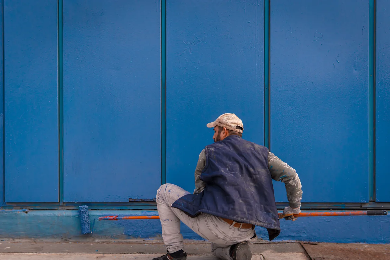 A painter rolling bold blue paint onto an outside wall in the sun