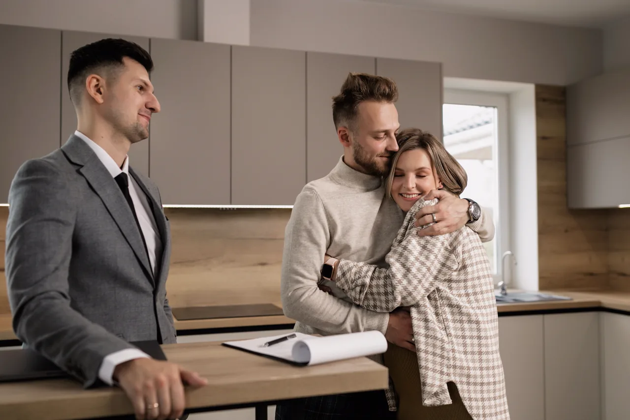 A real estate agent with a signed contract while a happy couple hug in their new kitchen